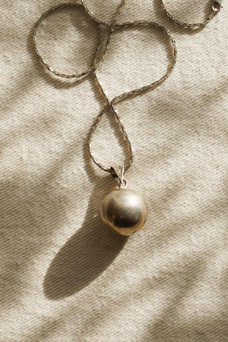Puede incluir: Un collar plateado con un colgante redondo. El colgante tiene una forma esf&eacute;rica lisa. La cadena tiene un dise&ntilde;o retorcido. El collar se muestra sobre una superficie clara y texturizada.