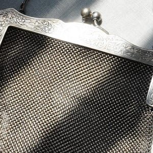 May include: A vintage silver mesh handbag with an ornate floral design on the frame. The bag features a chain handle and a clasp closure. The mesh is a dark silver color, and the frame is a lighter silver.