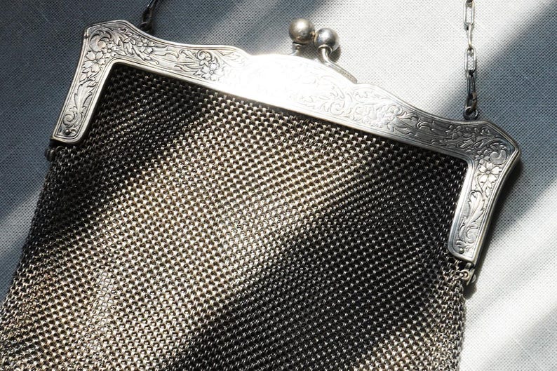 May include: A vintage silver mesh handbag with an ornate floral design on the frame. The bag features a chain handle and a clasp closure. The mesh is a dark silver color, and the frame is a lighter silver.