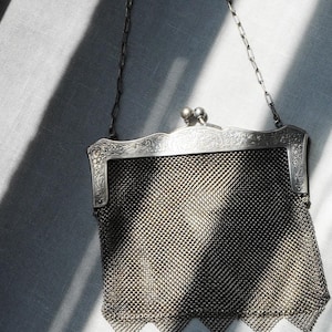 May include: An antique silver mesh purse with a decorative floral frame and a chain handle. The purse has a diamond-patterned mesh body and small dangling ornaments at the bottom. The clasp features two small, round embellishments.