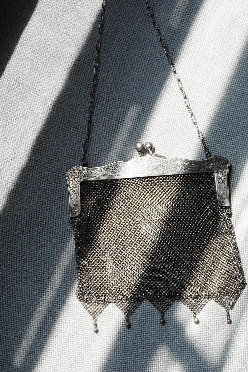 May include: An antique silver mesh purse with a decorative floral frame and a chain handle. The purse has a diamond-patterned mesh body and small dangling ornaments at the bottom. The clasp features two small, round embellishments.