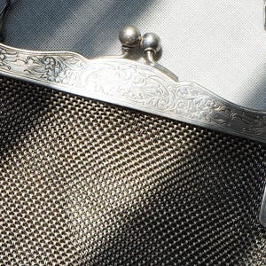 May include: Close-up of a vintage silver mesh purse. The purse features an ornate, floral-engraved frame and a delicate chain handle. The mesh body of the purse is a dark silver color, and the frame has a shiny finish.