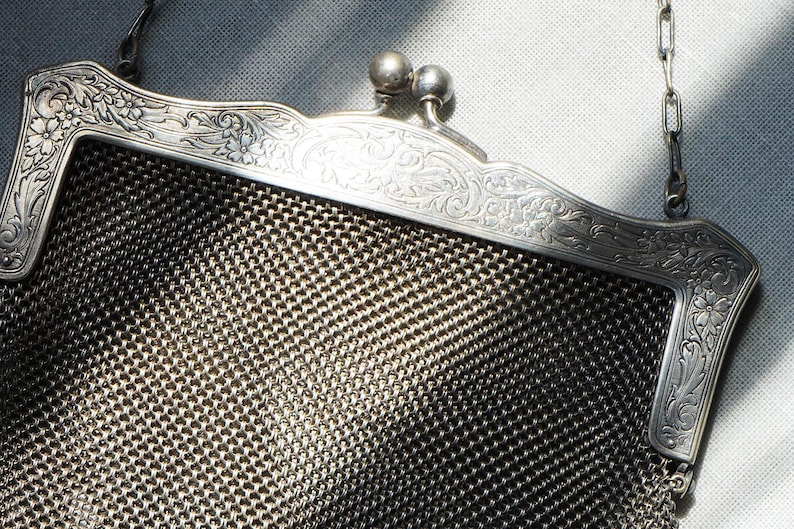 May include: Close-up of a vintage silver mesh purse. The purse features an ornate, floral-engraved frame and a delicate chain handle. The mesh body of the purse is a dark silver color, and the frame has a shiny finish.