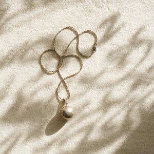 Puede incluir: Un collar de plata con un peque&ntilde;o colgante redondo. La cadena es fina y se encuentra en un patr&oacute;n curvo sobre una superficie texturizada de color crema. El colgante es una esfera met&aacute;lica lisa. La iluminaci&oacute;n crea sombras.