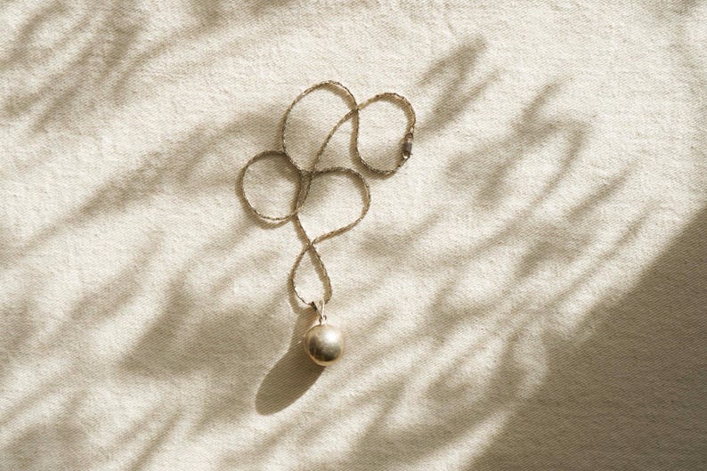Puede incluir: Un collar de plata con un peque&ntilde;o colgante redondo. La cadena es fina y se encuentra en un patr&oacute;n curvo sobre una superficie texturizada de color crema. El colgante es una esfera met&aacute;lica lisa. La iluminaci&oacute;n crea sombras.