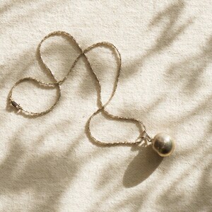 Puede incluir: Un collar plateado con una cadena fina y un colgante esf&eacute;rico. La joya descansa sobre una tela neutra, con sombras que a&ntilde;aden profundidad. El colgante mide aproximadamente 2 cm de di&aacute;metro. Una joya sencilla y elegante.