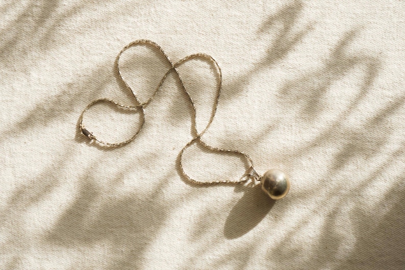 Puede incluir: Un collar plateado con una cadena fina y un colgante esf&eacute;rico. La joya descansa sobre una tela neutra, con sombras que a&ntilde;aden profundidad. El colgante mide aproximadamente 2 cm de di&aacute;metro. Una joya sencilla y elegante.