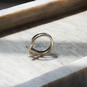 May include: A gold-colored earring with a looped design, resting on a marble surface. The earring features a double-banded loop with a small, decorative element at the bottom. The lighting creates shadows, highlighting the earring's shape.