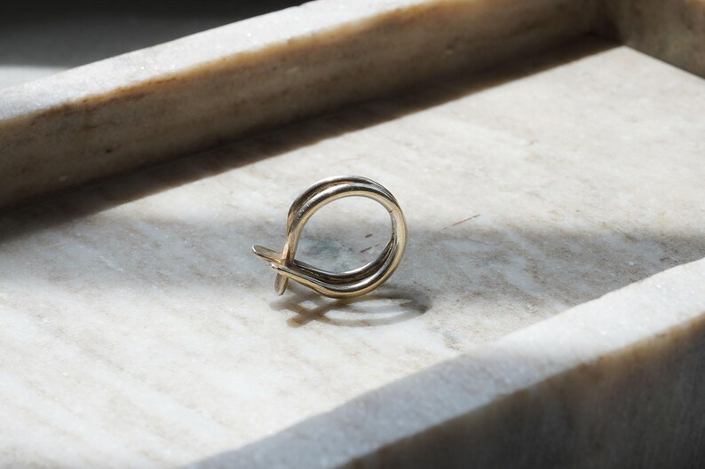 May include: A gold-colored earring with a looped design, resting on a marble surface. The earring features a double-banded loop with a small, decorative element at the bottom. The lighting creates shadows, highlighting the earring's shape.