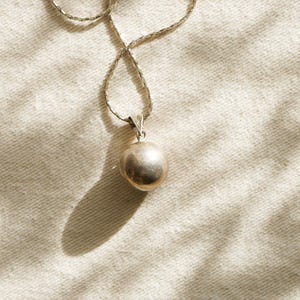 Puede incluir: Un collar con colgante plateado con un colgante redondo y liso. La cadena tiene un dise&ntilde;o tejido. El colgante cuelga sobre un fondo de tela texturizada blanquecina, con sombras.