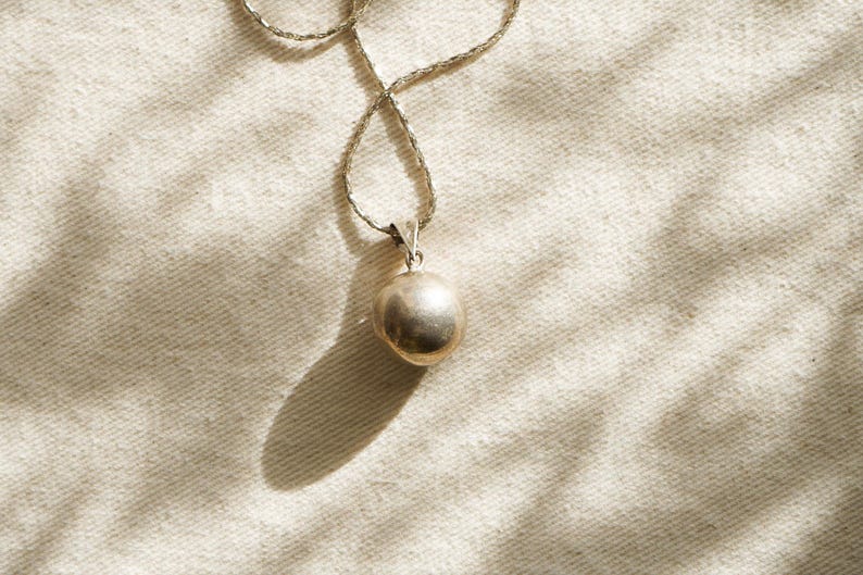 Puede incluir: Un collar con colgante plateado con un colgante redondo y liso. La cadena tiene un dise&ntilde;o tejido. El colgante cuelga sobre un fondo de tela texturizada blanquecina, con sombras.