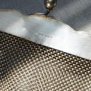 May include: Close-up of a vintage silver mesh purse. The purse features a decorative scalloped edge and a small ball clasp. The words "GERMAN SILVER" are stamped on the metal frame. The mesh is a tightly woven pattern.
