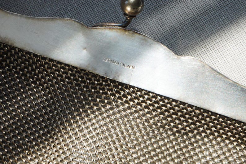 May include: Close-up of a vintage silver mesh purse. The purse features a decorative scalloped edge and a small ball clasp. The words "GERMAN SILVER" are stamped on the metal frame. The mesh is a tightly woven pattern.
