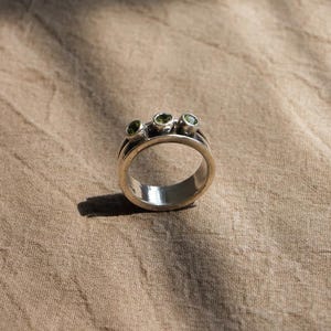 May include: A silver ring featuring a wide band with a spinning center section. The rotating band is set with three small, round, light green gemstones arranged in a horizontal row across the top surface of the metal ring structure.