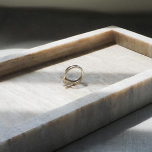 May include: A gold-colored ring with a double-band design rests inside a rectangular marble tray. The tray is white with gray veining and sits on a light-colored surface. The lighting creates shadows.