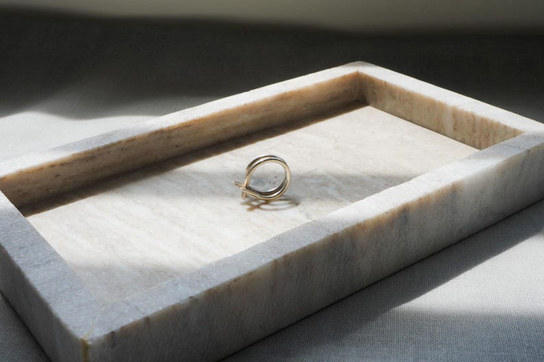 May include: A gold-colored ring with a double-band design rests inside a rectangular marble tray. The tray is white with gray veining and sits on a light-colored surface. The lighting creates shadows.