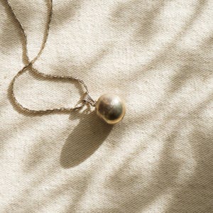 Puede incluir: Un collar de plata con un peque&ntilde;o colgante redondo de color dorado. La fina cadena descansa sobre una superficie texturizada de color crema. La sombra del colgante es visible, la escena est&aacute; iluminada por luz natural.