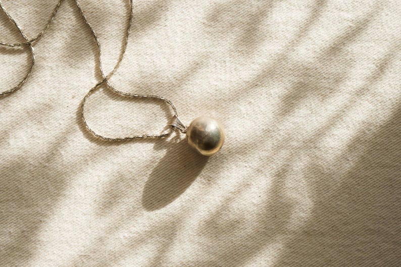Puede incluir: Un collar de plata con un peque&ntilde;o colgante redondo de color dorado. La fina cadena descansa sobre una superficie texturizada de color crema. La sombra del colgante es visible, la escena est&aacute; iluminada por luz natural.