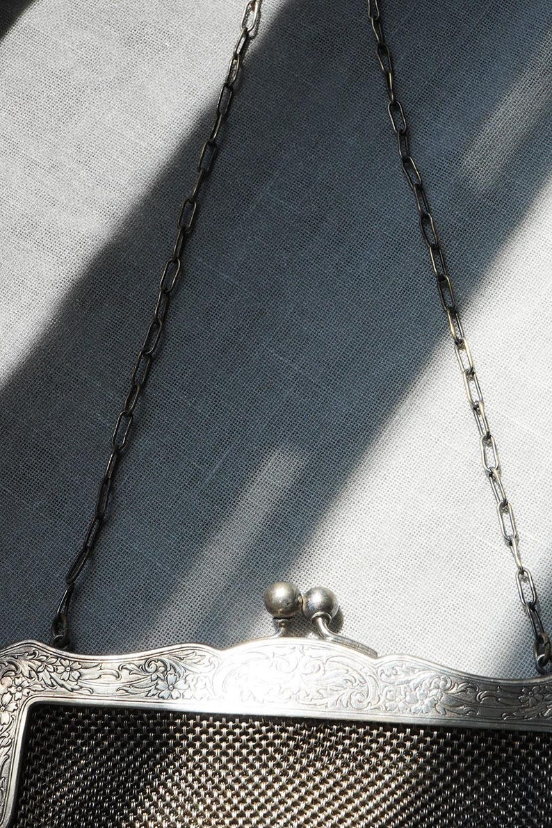 May include: Close-up of a vintage silver mesh purse with a decorative floral design on the frame. The purse has a chain handle and a clasp closure with two small, round silver balls. The mesh is a dark, woven pattern.