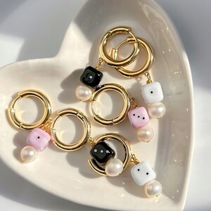 Può includere: Orecchini a cerchio dorati con ciondoli a forma di dado neri, bianchi e rosa, ciascuno abbinato a una perla. Gli orecchini sono esposti su un piatto bianco a forma di cuore, un accessorio giocoso ed elegante.