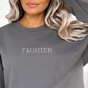 May include: A grey sweatshirt with the text "FIGHTER" and a ribbon symbol.