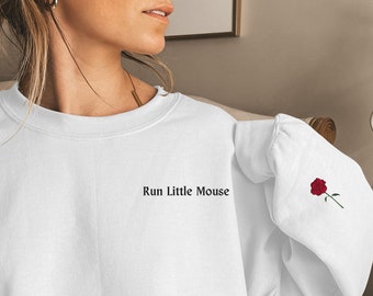 May include: White sweatshirt with the text "Run Little Mouse" in black on the front. A red rose with green stem and leaves is embroidered on the sleeve. The sweatshirt is a casual style.