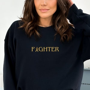 Puede incluir: Sudadera negra con una cinta dorada y la palabra "FIGHTER" impresa en la parte delantera.