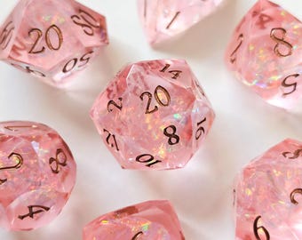 May include: A set of seven polyhedral dice made of translucent pink resin with iridescent flakes embedded inside. Each die features sharp edges and engraved numbers filled with a metallic copper-colored ink for high visibility.