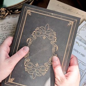 May include: A dark gray hardcover book featuring gold foil filigree detailing on the front cover. The cover design includes a central ornate frame with decorative corner accents and thin gold border lines surrounding the entire rectangular perimeter.
