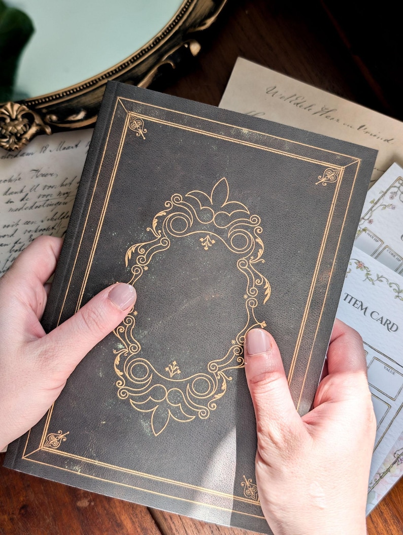 May include: A dark gray hardcover book featuring gold foil filigree detailing on the front cover. The cover design includes a central ornate frame with decorative corner accents and thin gold border lines surrounding the entire rectangular perimeter.