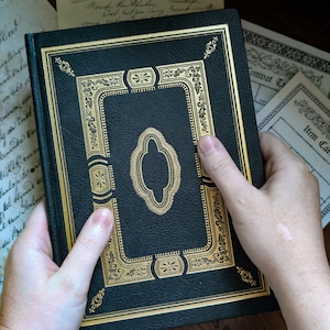 Puede incluir: Un libro de tapa dura negro con un intrincado estampado de l&aacute;mina dorada en la portada. El dise&ntilde;o incluye un marco decorativo central con patrones de filigrana ornamentados y bordes geom&eacute;tricos. La cubierta tiene un acabado texturizado.
