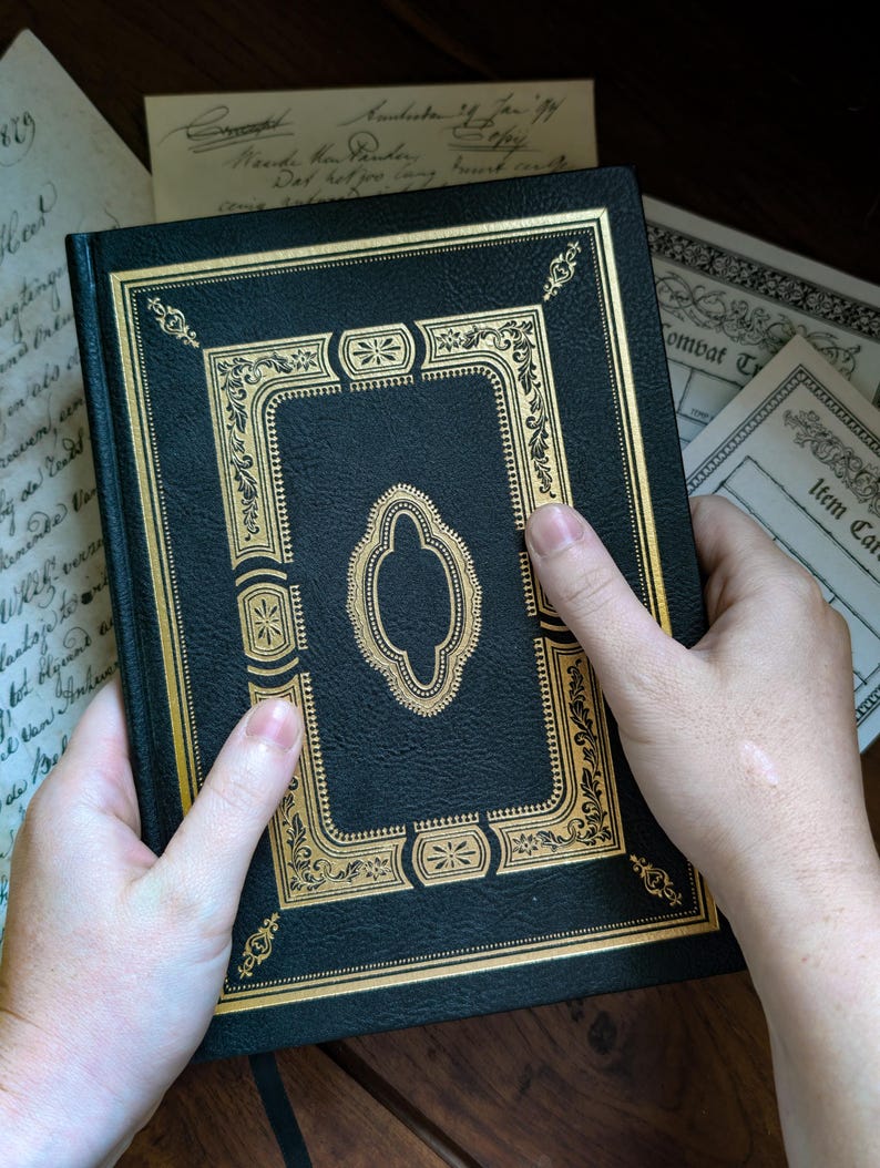 Puede incluir: Un libro de tapa dura negro con un intrincado estampado de l&aacute;mina dorada en la portada. El dise&ntilde;o incluye un marco decorativo central con patrones de filigrana ornamentados y bordes geom&eacute;tricos. La cubierta tiene un acabado texturizado.