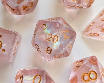 May include: A set of seven polyhedral dice made of translucent pale pink resin with an iridescent shimmer. Each die features engraved numbers filled with metallic gold ink, including a twenty-sided die centered in the arrangement.