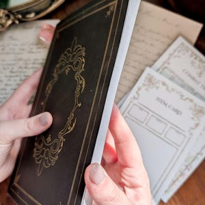 May include: A black notebook with gold foil filigree detailing on the cover, featuring a rectangular frame design. Beside the notebook are white paper cards with printed text including the words Item Card and Combat Tracker.