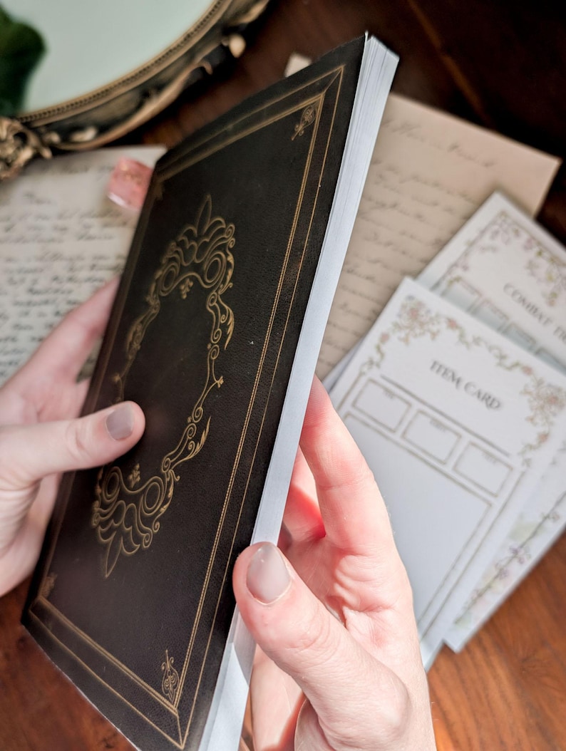 May include: A black notebook with gold foil filigree detailing on the cover, featuring a rectangular frame design. Beside the notebook are white paper cards with printed text including the words Item Card and Combat Tracker.