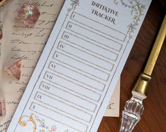 May include: A rectangular initiative tracker card featuring the text "INITIATIVE TRACKER" at the top, followed by ten numbered rows labeled I through X. The card is decorated with delicate floral illustrations and gold scrollwork.