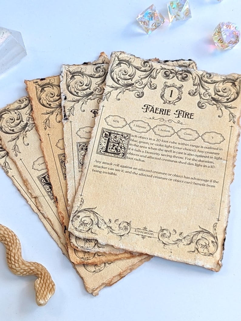 May include: A stack of aged paper cards with ornate black border designs and text. The top card reads "FAERIE FIRE" with game instructions. Several iridescent dice and a carved snake figurine are in the background.