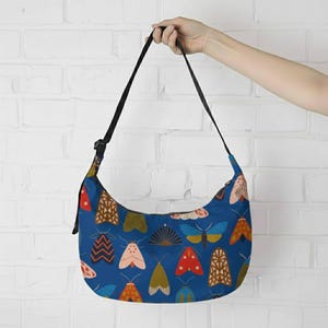 May include: A blue shoulder bag with a black strap and zipper. The bag is covered in a pattern of colorful moths in shades of red, yellow, brown, and pink. The bag is held up by a person's hand.