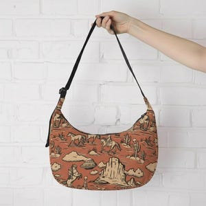 May include: A shoulder bag with a black strap and a southwestern-themed pattern. The pattern features horses, riders, cacti, clouds, and a UFO in shades of brown, tan, and black against a reddish-orange background.