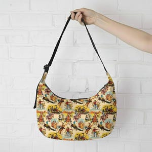 May include: A cream-colored shoulder bag with a black strap and a repeating pattern of cowboys and horses. The bag has a curved shape and a black adjustable strap.
