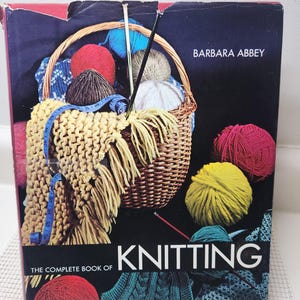 May include: A book titled "The Complete Book of Knitting" by Barbara Abbey. The cover features a wicker basket filled with yarn balls in various colors, including red, yellow, and blue. A knitted scarf and measuring tape are also visible.