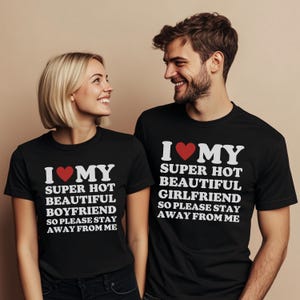 Peut inclure: Deux personnes portant des t-shirts noirs avec du texte blanc et un cœur rouge. Les chemises indiquent "I ❤️ MY SUPER HOT BEAUTIFUL BOYFRIEND SO PLEASE STAY AWAY FROM ME" et "I ❤️ MY SUPER HOT BEAUTIFUL GIRLFRIEND SO PLEASE STAY AWAY FROM ME".