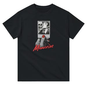 May include: Black t-shirt featuring a graphic of a black and white photo held by a hand with red nail polish. The photo shows a woman kissing a skull. The word "Memories" is written in red script below the hand.