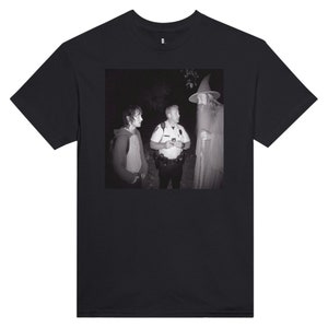 May include: Black t-shirt featuring a black and white image of a scene with three people: a police officer, a wizard, and a person in a hoodie. The image is a parody of a scene from a fantasy movie.