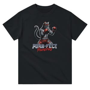 Puede incluir: Camiseta negra con un gato musculoso en atuendo de boxeo, incluyendo guantes y pantalones cortos rojos, y una cinta para la cabeza. El diseño incluye el texto "Purr-fect Punches" en una fuente estilizada.