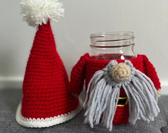Pode incluir: Um aquecedor de jarro de crochê de Papai Noel com barba branca e chapéu vermelho com pompom branco. O aquecedor de jarro está sobre uma superfície cinza.