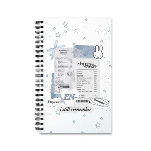 Peut inclure: Un cahier à spirales blanc avec un motif d'étoiles bleu clair. La couverture comprend du texte et des graphiques, tels qu'un design de reçu, une illustration de lapin et les mots "ROMANCE UNTOLD" et "ENHYPEN".