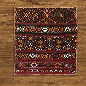 Puede incluir: Una alfombra rectangular estampada con un borde rojo oscuro y un diseño central con formas geométricas en negro, naranja, amarillo y blanco. La alfombra está colocada sobre una superficie de madera.