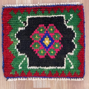 Puede incluir: Una pequeña alfombra cuadrada tejida a mano con un campo central negro. La alfombra presenta un diseño floral en rosa, azul, amarillo y verde. El borde es rojo con detalles verdes y blancos, y un borde azul. La alfombra tiene un borde con flecos.