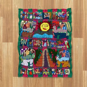 Könnte beinhalten: Ein farbenfrohes, besticktes Textil mit grünem Rand. Das zentrale Design zeigt Figuren, eine Sonne und eine Pyramide, alles in leuchtenden Farben wie Rot, Gelb, Blau und Braun. Das Kunstwerk ist wahrscheinlich von kultureller Bedeutung.